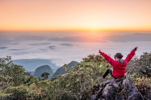 Mulher sentada e levanta as mãos na colina com neblina no nascer do sol no santuário de vida selvagem