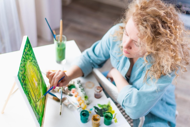 Mulher sentada em frente a uma pintura e pintando com pincéis sobre tela em casa