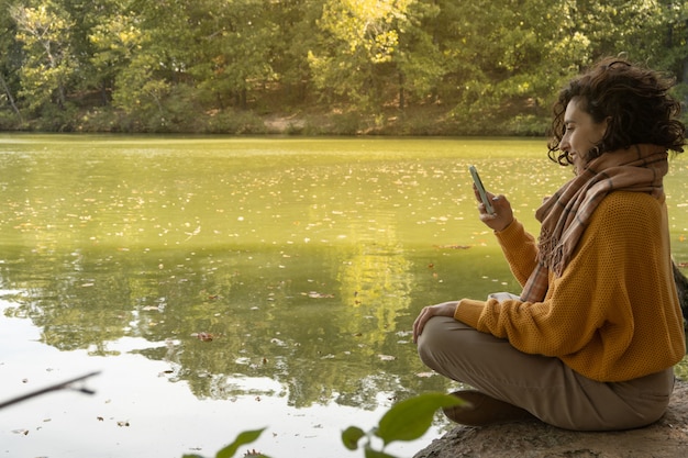 Foto mulher sentada em frente ao lago sorrindo enquanto olha para o celular com espaço de cópia