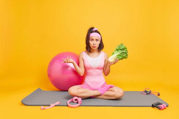 mulher sentada em posição de lótus no colchonete segura salada verde cercada por equipamentos esportivos mantém dieta saudável faz exercícios regulares fica em casa durante a quarentena