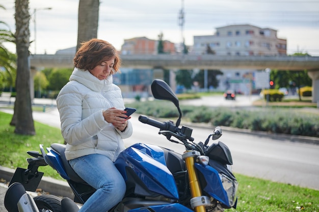 Mulher sentada em sua motocicleta estacionada, consulta seu celular