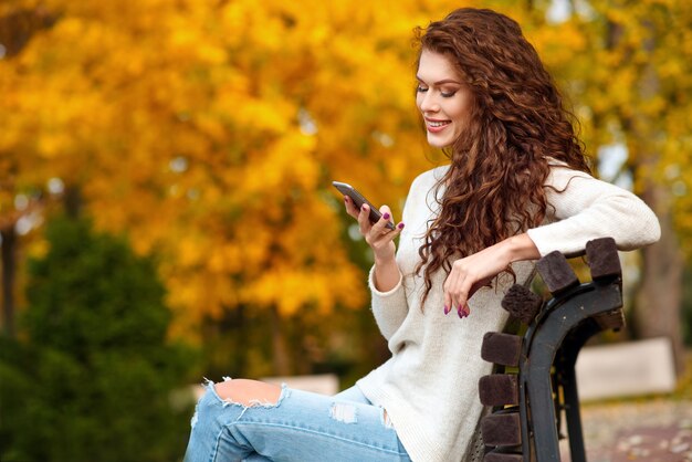 Mulher sentada em um banco no outono no parque segurando um telefone celular e sorrindo