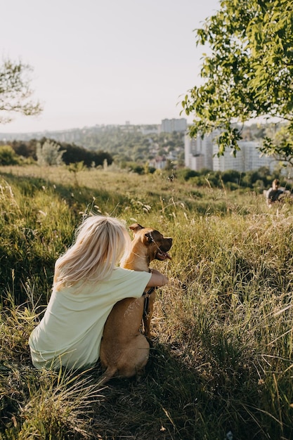 Mulher sentada em um prado com um cachorro abraçando-o olhando o pôr do sol