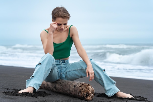 Mulher sentada em um tronco com as pernas abertas e a cabeça baixa, olhando para uma praia do Oceano Pacífico