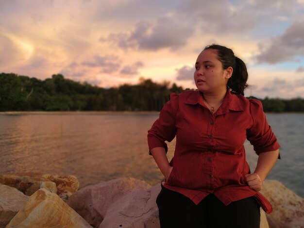 Foto mulher sentada em uma rocha ao lado do mar contra um céu nublado durante o pôr do sol