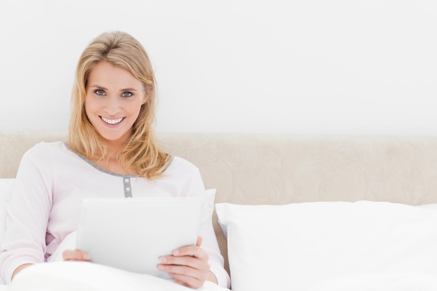 Mulher sentada na cama com tablet pc, olhando para a frente e sorrindo