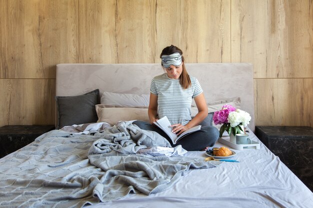 Mulher sentada na cama lendo um livro e tomando café da manhã.