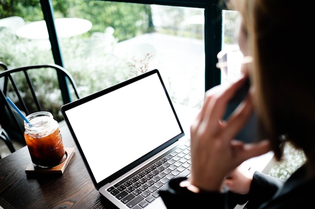 Mulher sentada na frente do laptop aberto com tela vazia em branco para suas informações ou conteúdo enquanto fala no telefone inteligente no café moderno