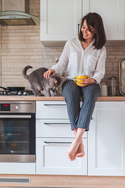 Mulher sentada na mesa da cozinha com gato bebendo chá da caneca amarela