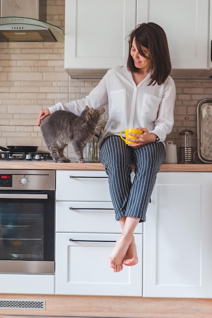 Mulher sentada na mesa da cozinha com gato bebendo chá da caneca amarela