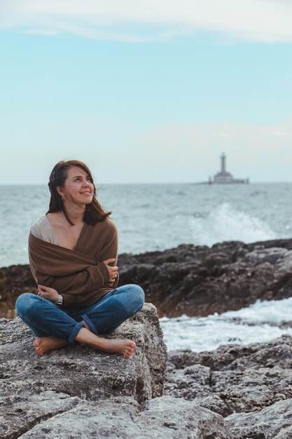 Mulher sentada na praia do mar rochoso no farol de jeans molhado no fundo do tempo ventoso férias de verão conceito despreocupado