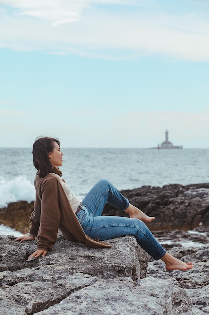 Mulher sentada na praia do mar rochoso no farol de jeans molhado no fundo do tempo ventoso férias de verão conceito despreocupado