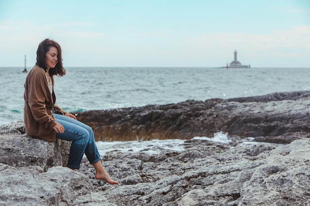 Mulher sentada na praia do mar rochoso no farol de jeans molhado no fundo