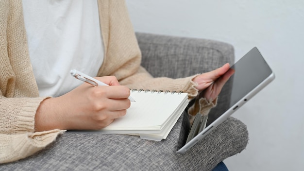 Mulher sentada na sala de estar usando tablet digital e escrevendo algo em seu bloco de notas em espiral