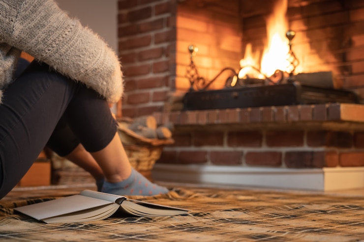 Foto mulher sentada no chão com um livro em frente ao calor da lareira no inverno.
