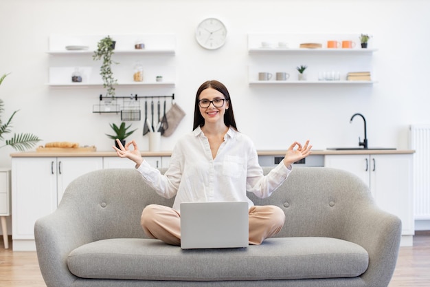 Mulher sentada no sofá com laptop sem fio na frente dela e meditando