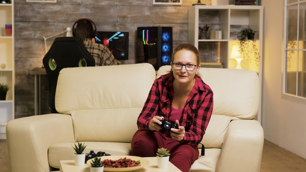 Mulher sentada no sofá na sala de estar jogando videogame usando controladores sem fio. Namorado jogando no computador em segundo plano.