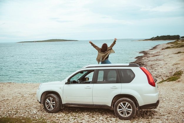 Mulher sentada no telhado do carro, apreciando a vista do mar. viagem de carro de férias de verão