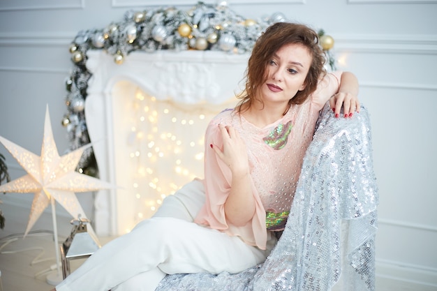 Mulher sentada perto da lareira na sala decorada. interior elegante de ano novo com árvore de natal, lareira branca, presentes e decorações. comemorando o natal