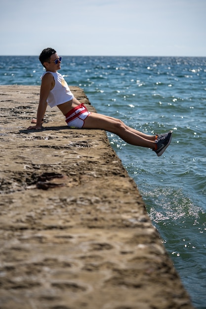 Mulher sentada sozinha no cais em shorts e camiseta com sapatos. Vista traseira. Mar de verão