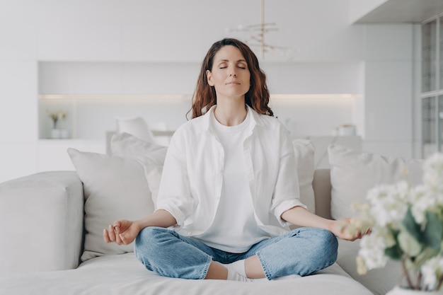 Mulher serena praticando meditação de ioga relaxante para alívio do estresse no sofá confortável em casa