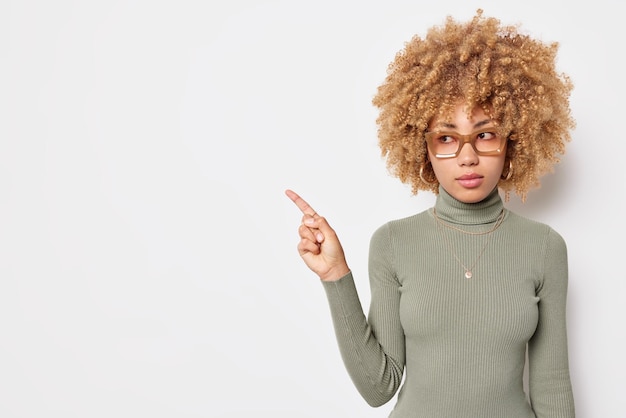 Mulher séria com cabelo loiro encaracolado mostra a bandeira de venda promocional de lado indica no espaço da cópia em branco usa óculos e poses de gola alta contra um fundo branco. Coloque seu anúncio aqui