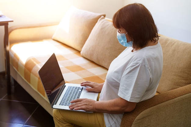 Mulher séria com máscara protetora usa laptop verifica e-mail na internet enquanto está sentado no sofá olhando ...