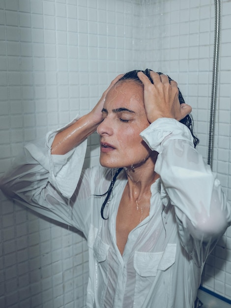 Mulher séria de camisa branca tocando o cabelo molhado enquanto toma banho na cabine e relaxa