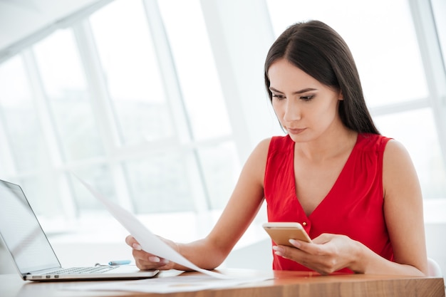 Foto mulher séria de camisa vermelha sentada à mesa com laptop, telefone e documentos no escritório