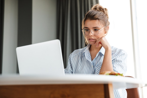 Mulher séria e simpática em óculos trabalhando com laptop enquanto está sentado à mesa na sala de estar