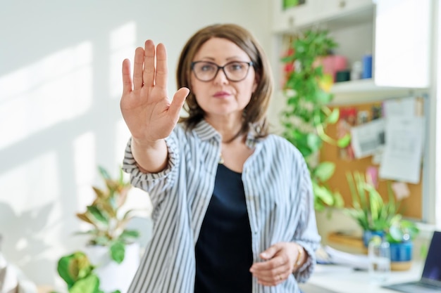 Foto mulher séria madura mostrando gesto de mão pare não no interior de casa sinal símbolo negação restrição proibição problema de controle