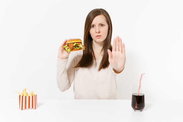 Mulher severa mostra gesto de pare na mesa com cola de batata frita de hambúrguer em frasco de vidro isolado no fundo branco. Nutrição adequada ou fast food clássico americano. Área de publicidade com espaço de cópia.