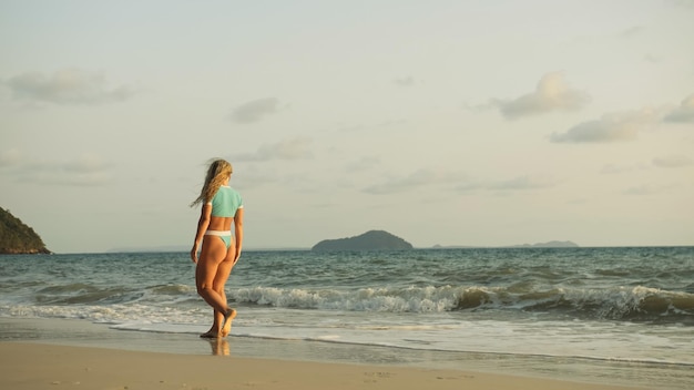 Mulher sexy andando sozinha no litoral da praia e relaxe o pôr do sol dourado quente Mulher andando na água em maiô azul e óculos de sol Conceito resto tropical resort viajando turismo feliz férias de verão