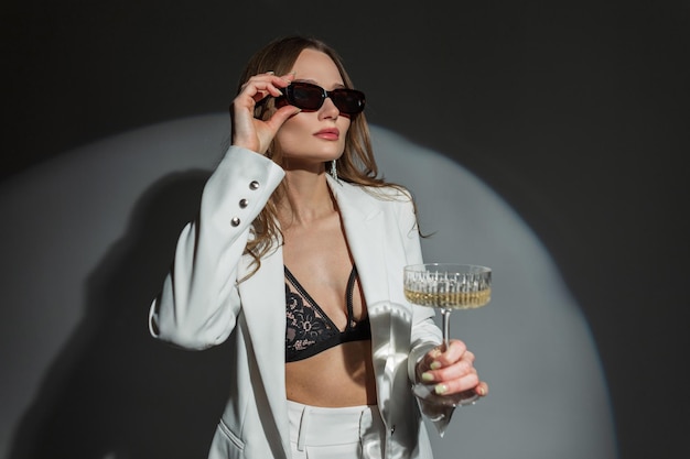 Mulher sexy bonita e bem-sucedida em terno branco da moda com blazer e sutiã de renda segura taça de champanhe e coloca óculos de sol em fundo escuro no estúdio Evento de celebração de senhora muito chique