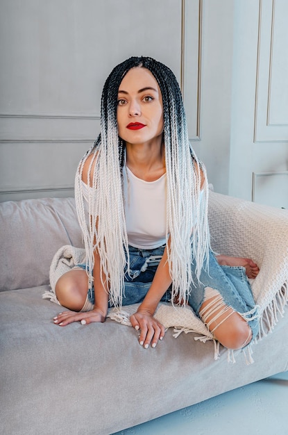 Mulher sexy com dreadlocks Camiseta branca e jeans azul Sentada em um sofá cinza de joelhos