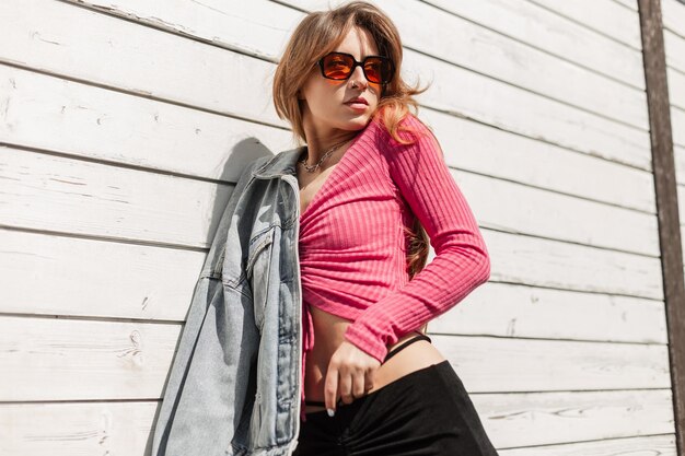 Mulher sexy e bonita moda com óculos de sol laranja legal vestindo roupa elegante e brilhante com uma jaqueta jeans cor-de-rosa e saia em pé perto de um edifício de madeira branco vintage