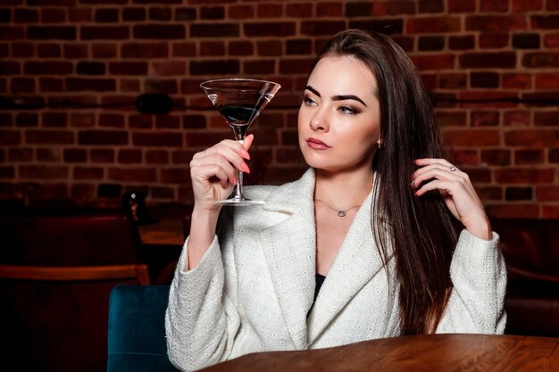 Mulher sexy elegante com copo de martini na mesa no restaurante de fundo interior à noite. Mulher de negócios morena encantadora está descansando em seu tempo livre. Conceito de fotografia publicitária. Copie o espaço