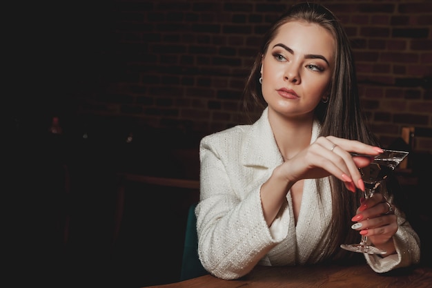 Foto mulher sexy elegante com copo de martini na mesa no restaurante de fundo interior à noite. mulher de negócios morena encantadora está descansando em seu tempo livre. conceito de fotografia publicitária. copie o espaço