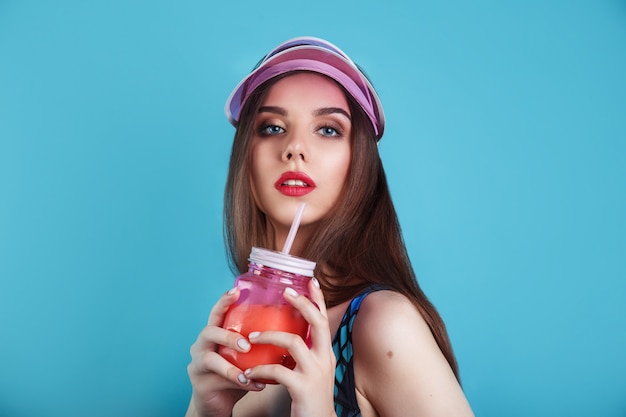 Mulher sexy maiô e viseira plástica, segurando o copo de bebida de verão fresco na parede azul