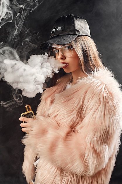 Mulher sexy na moda com casaco de pele rosa e boné fumando com cigarro eletrônico