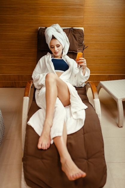 Mulher sexy no manto e toalha na cabeça relaxando com coquetel na cadeira do spa.