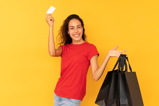 Mulher shopaholic alegre carregando sacolas de papel preto sobre fundo amarelo.