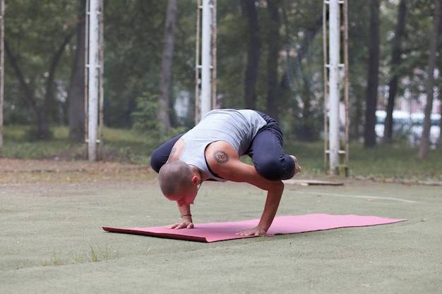 Mulher skinhead bronzeada com tatuagens étnicas fazendo exercícios de ioga - guindaste, ao ar livre em um parque verde