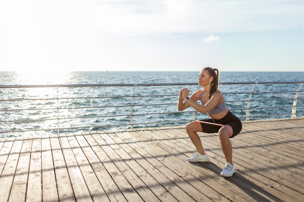 Mulher slim fit em roupas esportivas exercícios com elásticos de fitness na praia ao nascer do sol