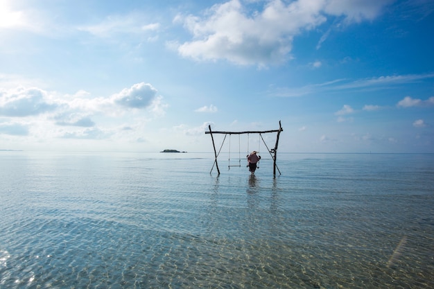 Mulher solitária balançando em um balanço de madeira sobre a água do mar rasa e calma com fundo de céu azul