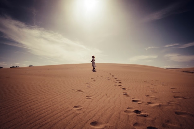 Mulher solitária caminhando livremente no deserto das dunas com um vestido branco elegante, desfrutando de atividades de lazer ao ar livre e um belo lugar ao redor - conceito de viagens e estilo de vida