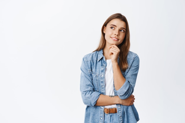 Foto mulher sonhadora e sorridente, imaginando ou pensando, olhando para o canto superior esquerdo e segurando a mão no queixo, lembra de algo interessante, tendo uma ideia, parede branca