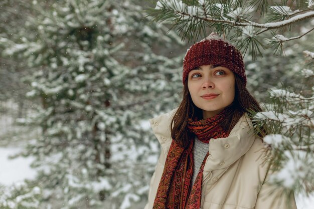 Mulher sonhadora em roupas quentes em pé debaixo de árvore nevada na floresta de inverno e desviar o olhar
