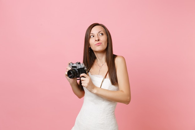 Mulher sonhadora em um vestido branco olhando para cima segurando uma câmera fotográfica vintage retrô escolhendo a equipe, o fotógrafo