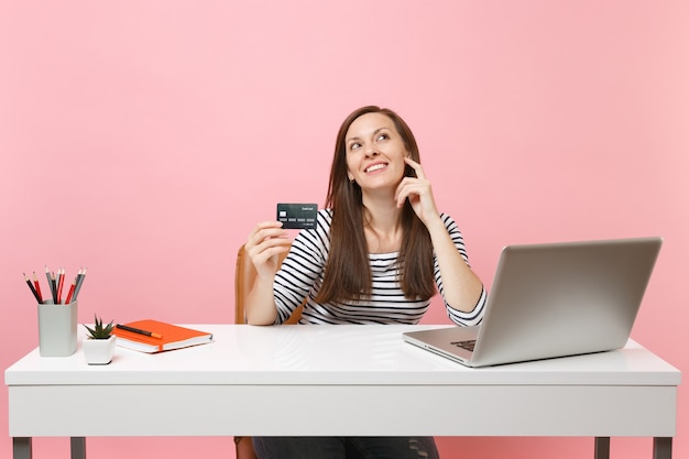 Mulher sonhadora, segurando o cartão de crédito, olhando para cima pensando em como gastar dinheiro enquanto trabalhava sentado no escritório com o laptop isolado em fundo rosa pastel. conceito de carreira empresarial de realização. copie o espaço.